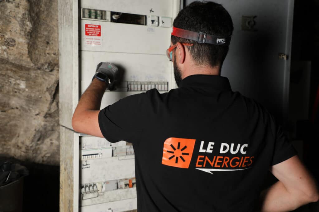 Électricien Le Duc Energies : Maintenance de tableau Électricien Le Duc Energies inspectant un tableau électrique muni d'une lampe frontale et de lunettes de sécurité.