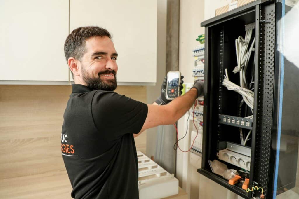 Électricien Luc Energies : Installation de Tableau Électrique Électricien Luc Energies souriant utilisant un multimètre sur un tableau électrique pour une installation sécurisée.