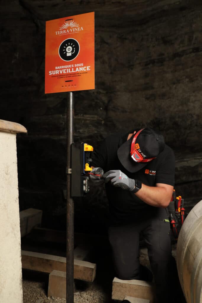 Terra Vinea : Installation alarme par Le Duc Énergie Technicien Le Duc Énergie installant une alarme Terra Vinea sous un panneau Barriques sous surveillance.