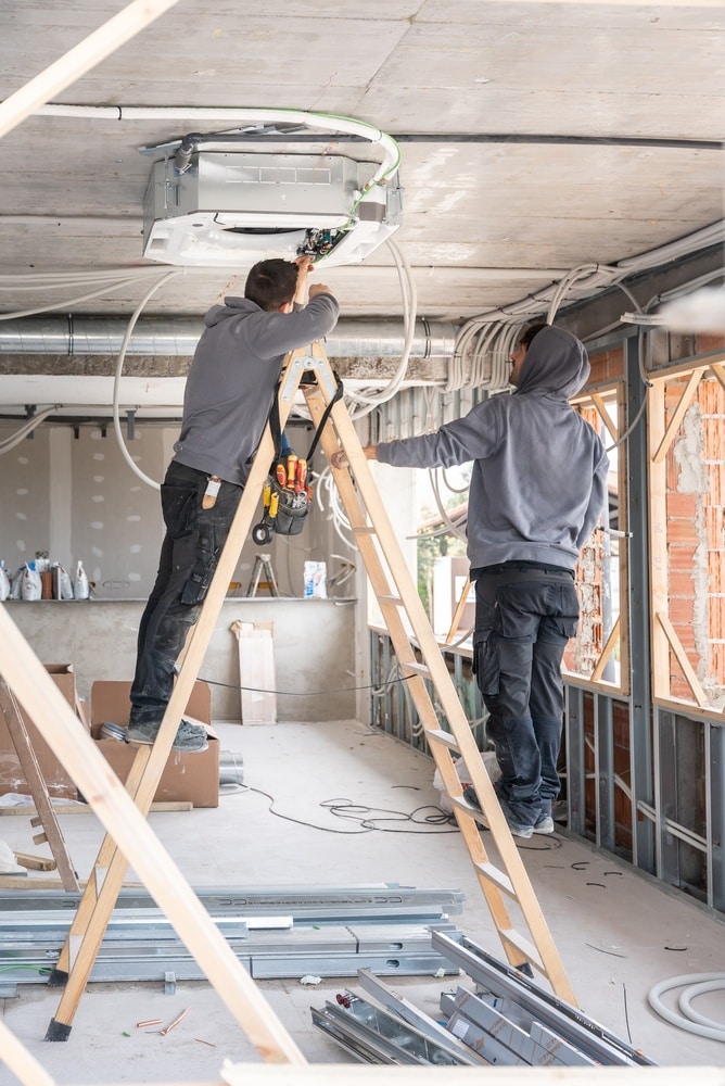 Installation Climatisation : Professionnels sur Chantier Deux techniciens installent un climatiseur au plafond sur un chantier de rénovation à l'aide d'une échelle en bois.