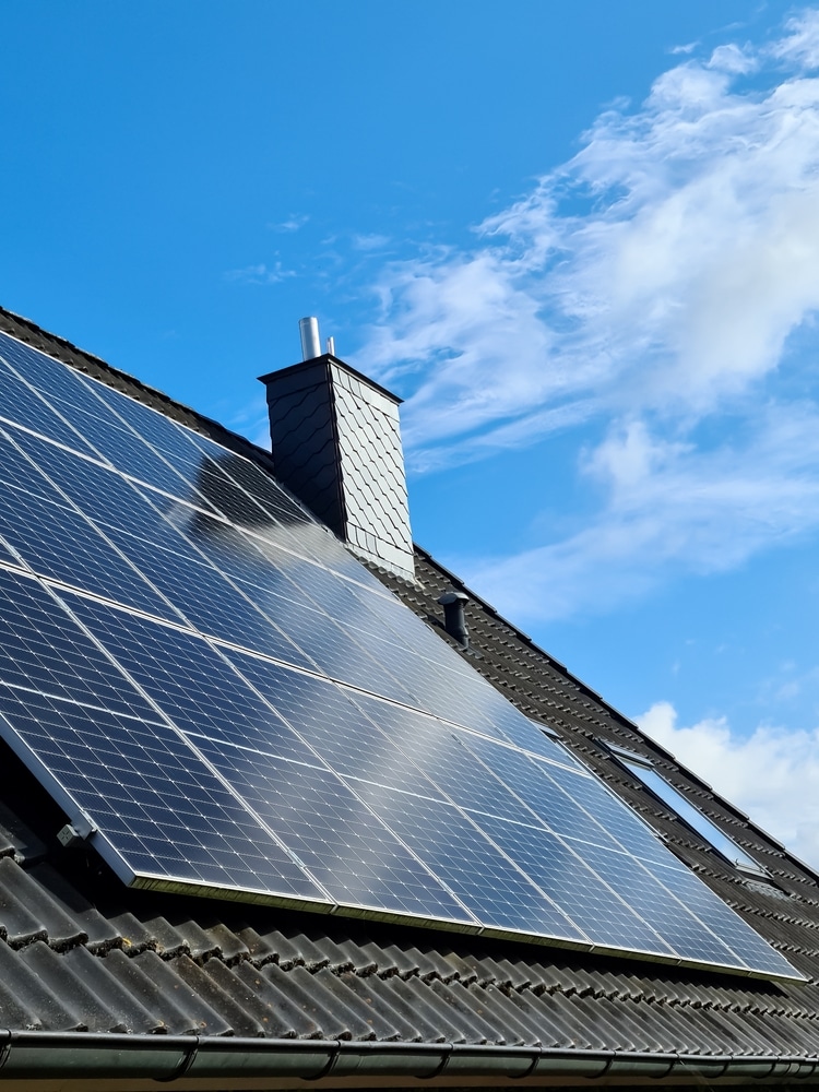 Installation de Panneaux Solaires sur un Toit de Maison Panneaux solaires installés sur un toit en tuiles sombres sous un ciel bleu avec une cheminée.