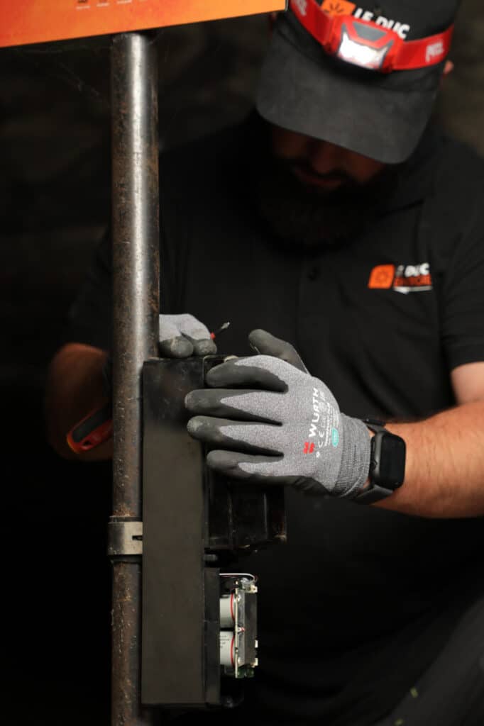Maintenance électrique experte par un technicien Le Duc Technicien Le Duc en gants Würth réparant un boîtier électronique sur un mât avec un tournevis et lampe frontale.